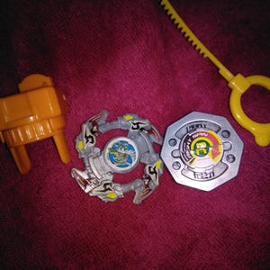 May include: A selection of toy parts, including a yellow launcher, a silver and grey spinning top, and a yellow ripcord. The spinning top features a detailed design with a central image. The items are arranged on a pink surface.