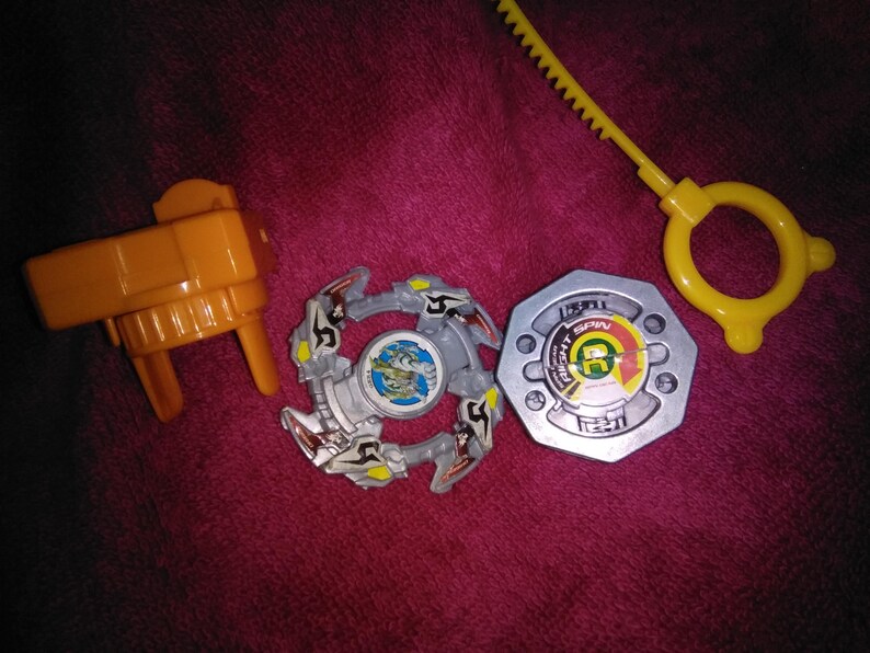 May include: A selection of toy parts, including a yellow launcher, a silver and grey spinning top, and a yellow ripcord. The spinning top features a detailed design with a central image. The items are arranged on a pink surface.