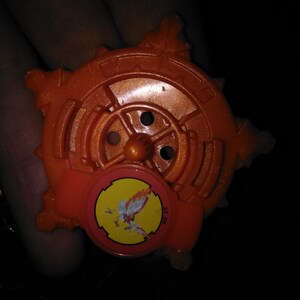 Pode incluir: Brinquedo de pl&aacute;stico laranja com um design circular e um pequeno emblema redondo com uma criatura semelhante a um p&aacute;ssaro. O brinquedo tem uma apar&ecirc;ncia de engrenagem com m&uacute;ltiplas pontas e um bot&atilde;o central. O emblema &eacute; amarelo com um p&aacute;ssaro vermelho e branco.