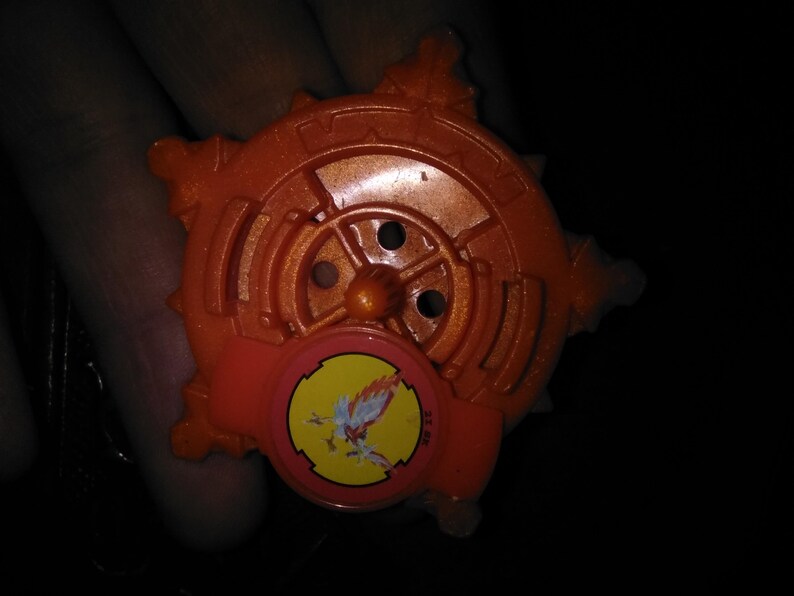 Pode incluir: Brinquedo de pl&aacute;stico laranja com um design circular e um pequeno emblema redondo com uma criatura semelhante a um p&aacute;ssaro. O brinquedo tem uma apar&ecirc;ncia de engrenagem com m&uacute;ltiplas pontas e um bot&atilde;o central. O emblema &eacute; amarelo com um p&aacute;ssaro vermelho e branco.