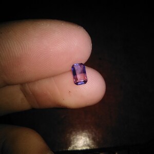 May include: A small, rectangular-cut gemstone featuring a vibrant gradient of purple and pink hues. The stone is faceted, allowing it to catch and reflect light, displaying a clear, transparent quality with distinct colour transitions across its surface.