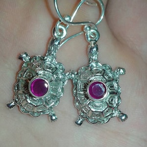 Puede incluir: Pendientes plateados con forma de tortuga y una gema de color magenta en el centro. Los pendientes tienen un diseño calado detallado y están suspendidos de un gancho simple. Una joya única.