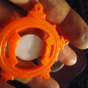 以下が含まれることがあります： オレンジ色のプラスチック製のオブジェクトのクローズアップ。円形のデザインで、中央は白。オブジェクトには、一連の尖ったエッジと小さな装飾的な突起があります。「HUDSON SOFT」の文字が見えます。手で持っています。