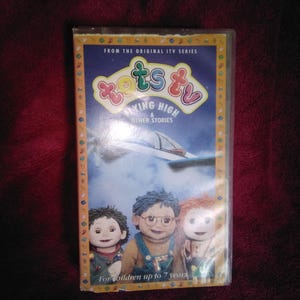 May include: A VHS video cassette for the children's show Tots TV, titled "Flying High & Other Stories." The cover shows three doll-like characters and the text "For children up to 7 years."