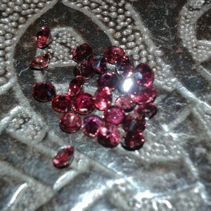 Può includere: Un gruppo di piccole gemme rotonde e sfaccettate in varie tonalità di rosso e rosa. Le gemme sono sparse su una superficie argentata e strutturata, catturando la luce. Adatto per la creazione di gioielli.