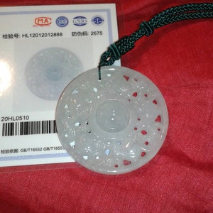 May include: A pale green jade pendant with intricate floral carvings, suspended from a dark green cord. The pendant is circular with a central design and openwork details. A certificate with text is visible.