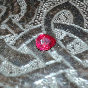 May include: A faceted, triangular-shaped, red gemstone. The gem is set against a textured, silver-colored surface with a repeating pattern. The gemstone is a vibrant, deep red color.