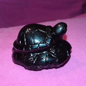 Puede incluir: Una figura de tortuga negra brillante con un caparazón y una cabeza detallados, posada sobre una base texturizada. La figura se encuentra sobre un fondo rosa, mostrando su superficie pulida y su intrincado diseño.