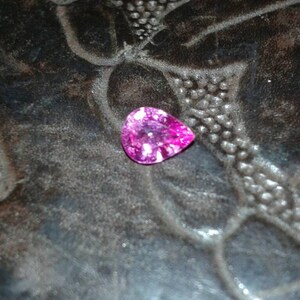 May include: A pear-shaped, bright pink gemstone with white markings. The gem is displayed on a dark, textured surface, providing a strong contrast. The gemstone's cut reflects light, highlighting its sparkle.