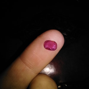 May include: A small, oval-shaped, deep pink gemstone. The gem is held on a finger against a dark background, highlighting its color and shape. The gem appears to be a ruby, showcasing its natural beauty and potential for jewelry.