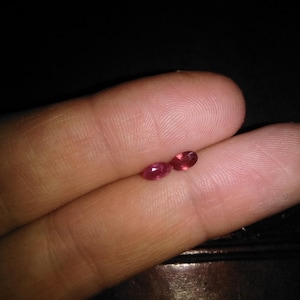 Peut inclure: Deux petites pierres précieuses rouges de forme ovale sont présentées sur les doigts d'une personne. Les pierres précieuses sont d'une teinte rouge vif et leurs surfaces polies reflètent la lumière. Le fond est sombre.