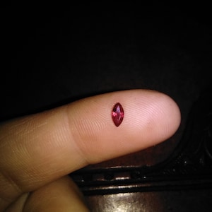 Puede incluir: Una pequeña gema roja con corte marquesa se muestra en la punta de un dedo sobre un fondo oscuro. La gema tiene forma ovalada puntiaguda y un color rojo intenso y vibrante, que refleja la luz. La imagen destaca el tamaño y la claridad de la gema.