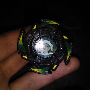 May include: A black and green spinning top toy with yellow accents. The center features a circular image of a green dragon. The toy has the text "M-DRAGON" and "4763" printed on it.
