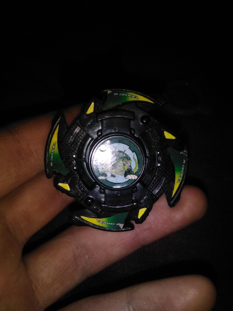 May include: A black and green spinning top toy with yellow accents. The center features a circular image of a green dragon. The toy has the text "M-DRAGON" and "4763" printed on it.