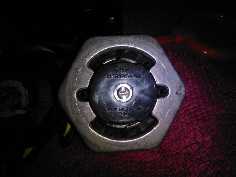 May include: Close-up of a silver and black hexagonal object. The object has a central black circular component with a silver emblem and the text "&copy;2002 HASBRO, INC. CHINA". The text "OH/T TAKARA 02" is also visible.