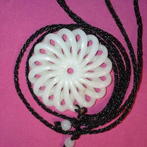 Op de afbeelding: Witte hangerketting met een bloemvormig ontwerp. De hanger heeft een gebeeldhouwde, cirkelvormige vorm met opengewerkte details, hangend aan een zwart koord. De ketting wordt gepresenteerd tegen een roze achtergrond.