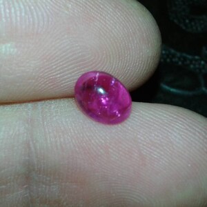 Puede incluir: Una vibrante gema de rubí de forma ovalada. La gema es de un color rojo rosado profundo y saturado, con una superficie pulida y reflectante. El rubí se sostiene entre dos dedos, mostrando su tamaño y brillo. Una vista de cerca resalta la claridad de la gema.
