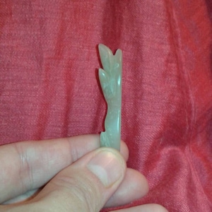 May include: A light green, carved jade figurine. The jade is held in a hand and has a stylized leaf or plant design. The background is a red fabric.