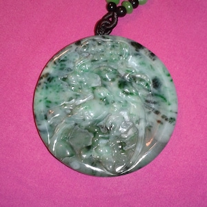 May include: A circular, green jade pendant featuring carved figures, hanging from a black cord necklace with green, yellow, and black beads. The pendant has a mottled appearance with darker green spots.