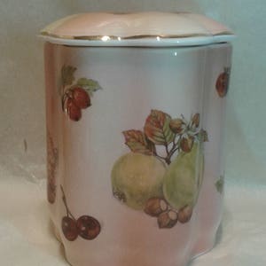 May include: A ceramic jar with a lid, featuring a light pink background and fruit illustrations. The jar has a gold trim and is decorated with images of pears, cherries, and nuts. The lid is shaped like a flower.