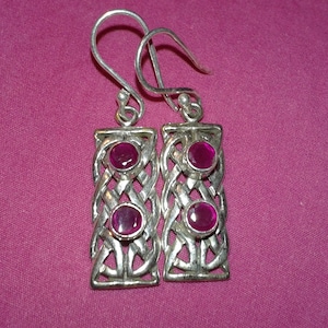 Puede incluir: Un par de pendientes plateados con un diseño rectangular. Cada pendiente presenta tres gemas redondas de color rosa intenso engastadas en un patrón abierto y tejido. Los pendientes cuelgan de sencillos ganchos para las orejas sobre un fondo rosa.