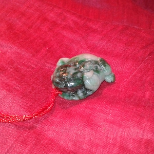 May include: A jade pendant carved in the shape of a small frog. The frog is green and white with a red cord attached. The pendant is set against a red background.