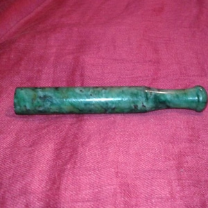 May include: A green jade pipe with a cylindrical body and a flared mouthpiece. The jade has dark green and black marbling. The pipe is displayed on a red fabric surface.