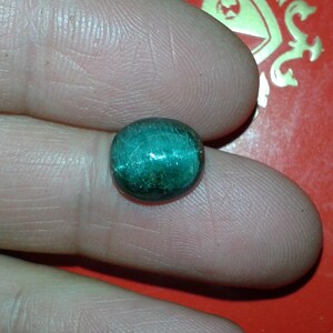 May include: A round, polished, teal gemstone. The stone has a reflective, shimmering quality, with visible internal striations. The gem is held between two fingers against a red background, suggesting a jewelry or craft item.