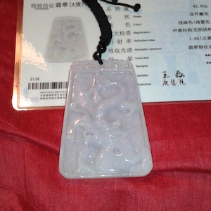 Puede incluir: Colgante rectangular de jade con un diseño tallado, suspendido de un cordón negro. El colgante es de color verde pálido y púrpura. Un certificado con texto chino está en el fondo.