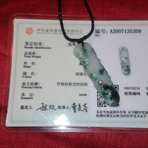 May include: A jade pendant with a black cord, displayed on a white card with Chinese text. The pendant is a light green color with darker green spots. The card includes a QR code and a serial number.