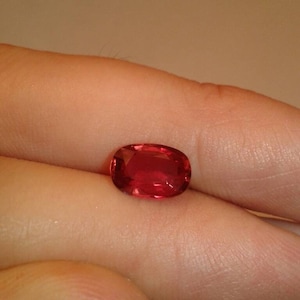 May include: A vibrant, oval-cut, red gemstone. The gem is held in a hand, showcasing its polished surface and rich color. The stone's facets catch the light, creating a sparkling effect. This gemstone is a beautiful example of fine jewelry.