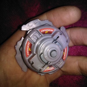 May include: A grey and red Beyblade toy, with the text "Driger F" in red. The toy has a circular design with multiple sections and a central metal piece. The image is a close-up shot of the toy.