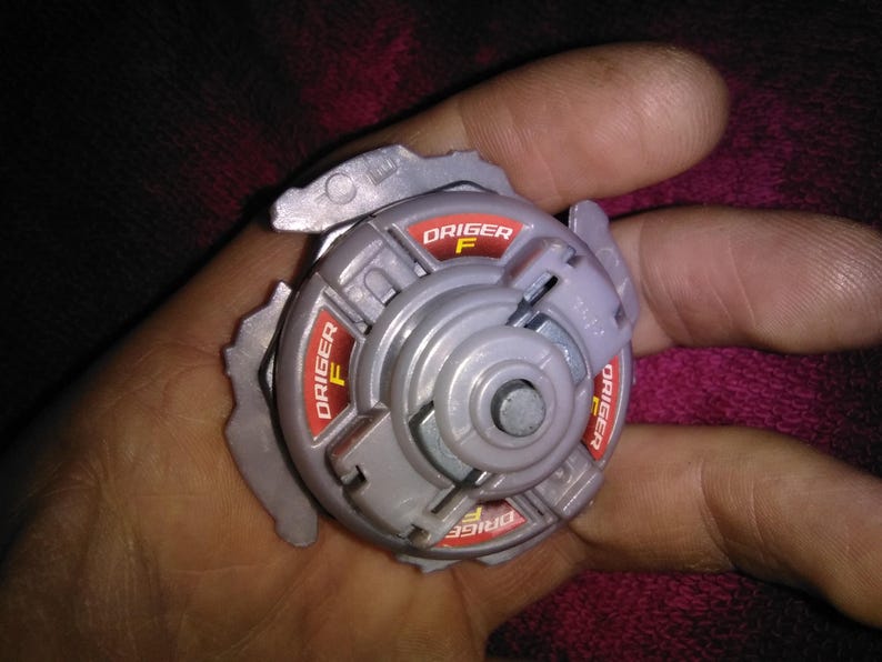 May include: A grey and red Beyblade toy, with the text "Driger F" in red. The toy has a circular design with multiple sections and a central metal piece. The image is a close-up shot of the toy.