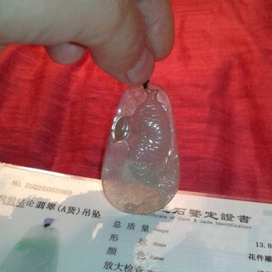 May include: A translucent, teardrop-shaped jade pendant with a carved design, held by a hand. The pendant is light green and white, with a black cord. A certificate with Chinese characters is visible.