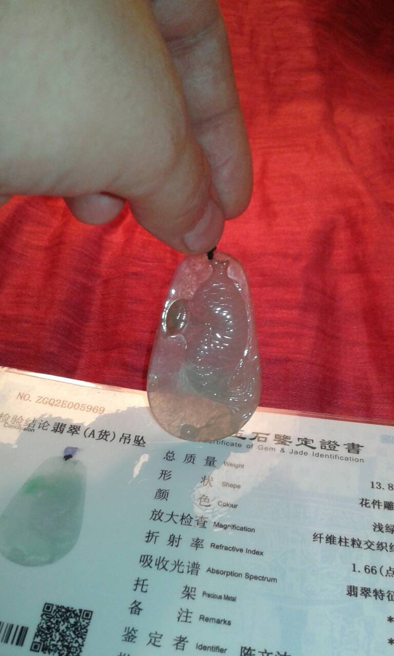 May include: A translucent, teardrop-shaped jade pendant with a carved design, held by a hand. The pendant is light green and white, with a black cord. A certificate with Chinese characters is visible.