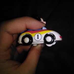 May include: A small, white toy race car with black wheels and the number 1 on the side. The car has a red, yellow, and blue stripe design. A small figure is seated in the car.