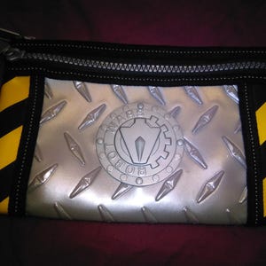 May include: A rectangular pouch with a silver diamond plate design and a black zip. The pouch has yellow and black diagonal stripes on the sides. The centre features a circular emblem with the words "Robot Wars".