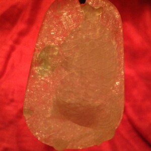 May include: A translucent, pendant-style jade carving, featuring a detailed relief of a fish. The pendant is suspended from a black cord. The jade has a light, almost white color, with subtle variations. The background is a solid red.