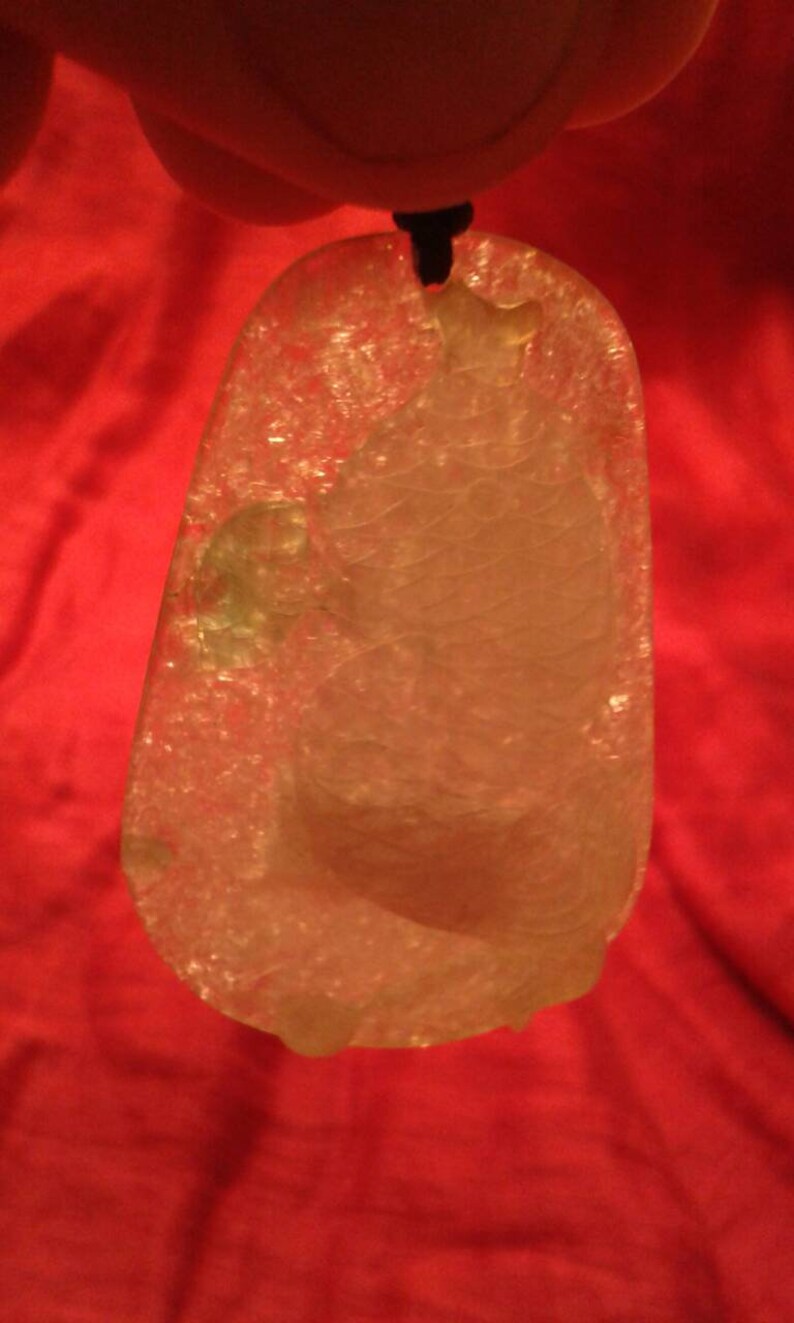 May include: A translucent, pendant-style jade carving, featuring a detailed relief of a fish. The pendant is suspended from a black cord. The jade has a light, almost white color, with subtle variations. The background is a solid red.