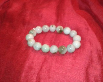 Puede incluir: Un brazalete de cuentas redondas y claras, con sutiles acentos verdes y marrones. El brazalete se exhibe sobre un fondo rojo vibrante, mostrando sus tonos y texturas naturales.