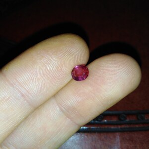 Könnte beinhalten: Ein kleiner, runder, facettierter Rubin-Edelstein. Der Edelstein hat einen leuchtenden Rotton mit einem leichten Rosa-Stich. Der Rubin wird zwischen zwei Fingern gehalten, um seine Größe und Farbe zu zeigen.