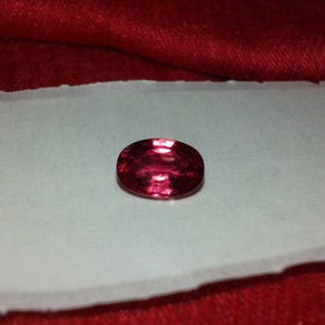 May include: A faceted, oval-shaped, deep red gemstone rests on a white surface against a red background. The gem's polished surface reflects light, showcasing its internal structure and color variations. This gemstone is likely a ruby or garnet.
