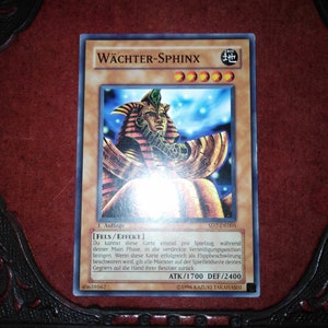 May include: A Yu-Gi-Oh! trading card featuring the "Wächter-Sphinx" monster. The card has a gold border and a detailed illustration of a sphinx with a pharaoh headdress. The card's text is in German, with ATK/1700 and DEF/2400 stats.