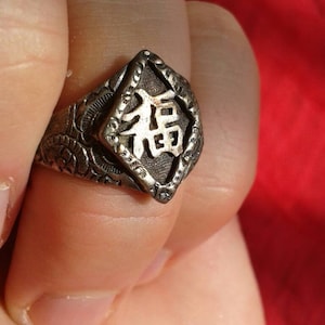 Può includere: Anello d'argento con un design in rilievo e un elemento centrale a forma di diamante. L'elemento centrale presenta un carattere cinese. L'anello è tenuto tra le dita su uno sfondo rosso.