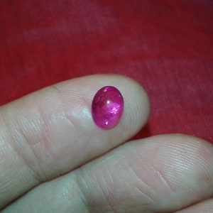 May include: A vibrant, oval-shaped, pink gemstone. The polished stone is held between two fingers against a red background. The gem has a glossy surface and appears to be translucent, with internal light reflections.
