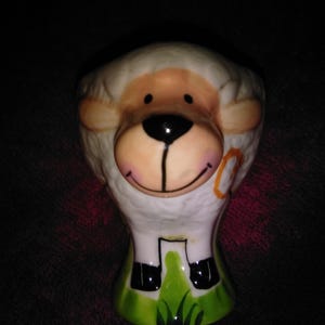 Op de afbeelding: Een keramische eierdop in de vorm van een schaap. De eierdop heeft een witte body met een schapengezicht, inclusief een zwarte neus, een lachende mond en beige oren. De basis is groen met gras en zwarte hoeven.