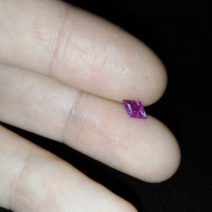 May include: A small, square-cut, vibrant magenta gemstone. The gem is held between two fingers against a dark background, showcasing its facets and colour. The gem's shape is a modified square or rectangle.