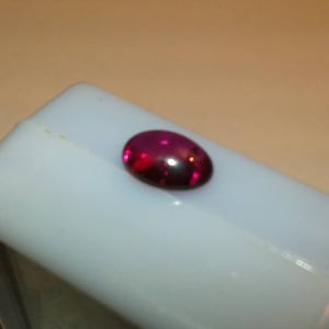 May include: A polished, oval-shaped, deep red gemstone rests on a light blue surface. The gemstone has a glossy finish, reflecting light. The setting is a simple, rectangular shape, suggesting a display or storage container for the gem.