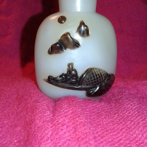 May include: A white, oval-shaped glass bottle with a matching stopper. The bottle features a carved scene of a person in a boat, with mountains and other details in brown. The bottle is displayed on a pink surface.