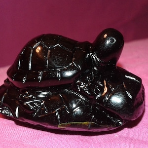 Puede incluir: Figura de tortuga negra brillante con detalles en el caparazón y la cabeza. La tortuga descansa sobre una base texturizada, creando un elemento decorativo. La figura está sobre un fondo rosa.